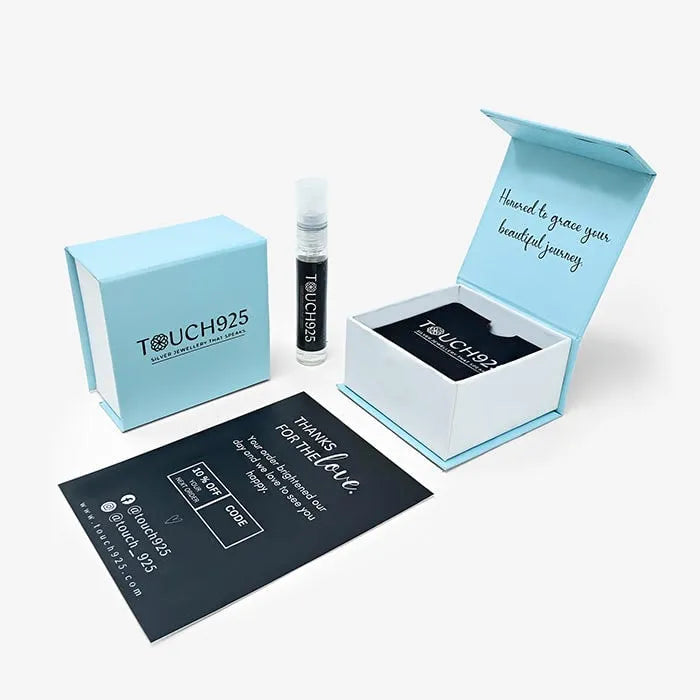 A light blue gift box with "Touch 925" printed on the front is open, revealing a black jewelry pouch containing a stunning Majestic Roar Zircon Studded Rose Gold Locket. Beside it is a small spray bottle labeled "Touch 925." A thank-you card with social media handles and a message is placed next to the items.